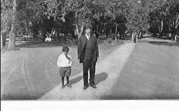 [Here's a pleasant photo of Robert M. Wright and his son Connor taking a stroll along a tree-lined dirt path in Wright Park, Dodge City, KS. Robert Wright is dressed much as he was in the formal photograph presented a few pages back, but somehow manages to look much more casual and even a bit animated. The son is decked out in shoes, knee-length socks reaching up to beneath what are either knee-length pants or knickers, a white shirt, the tails of which are struggling to escape from the clutches of the pants and hang out in a more natural fashion. In addition, the son has been provided with a necktie. If the boy's trousers are indeed knickers, his outfit is not unlike the ensemble that I wore as a child. Shirtails seem not to have changed much.]