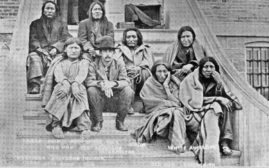 [Photograph] (somewhat the worse for wear)of a group of Indians. Seven Indians, clad in blankets or rough coats, are sitting on a flight of steps and surrounding a youngish Whiteface. The latter is slim, with a clean-looking face embellished by a simple straight moustache. His trousers are neatly tucked into the top of his fine-looking boots and he is wearing his black hat at a slight but jaunty angle. There is a set about his face and eyes that suggests that he would not be the sort of man you would like to cross. The faces of the Indians display a mixture of emotions - boredom, confusion, anger, all with more than a touch of desperation.]